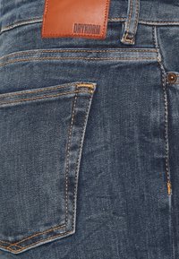 Pantalones vaqueros de mezclilla azul oscuro con costuras naranjas, un bolsillo trasero con una leve curvatura y una etiqueta de cuero marrón con el lema "DRYKORN" grabado.