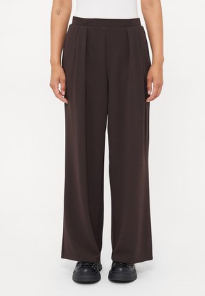 Persona vistiendo pantalones de tiro alto y pierna ancha de color marrón oscuro combinados con zapatos negros de cordones, de pie contra un fondo blanco.