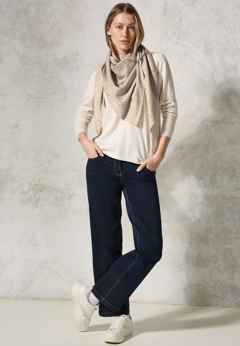 Sciarpa triangolare beige con piccoli pois drappeggiata su una maglietta a maniche lunghe crema, abbinata a jeans dritti scuri e sneakers bianche.