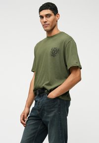 T-shirt de algodão verde-azeitona com mangas curtas, apresentando um emblema preto no peito e um pequeno logótipo na manga, combinado com jeans escuros.