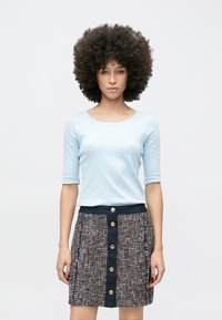 Lichtblauw aangesloten topje met driekwart mouwen, gecombineerd met een getextureerde rok met een veelkleurige stof en gouden knopen aan de voorkant.