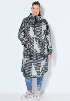 Kvinde smilende, iført en lang sort-hvid mønstret hættetrøje, blå leggings og grå sportssko, stående mod en ensfarvet hvid baggrund.