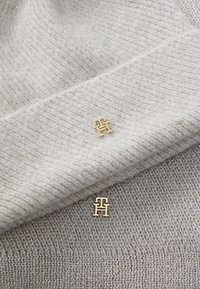 Lichtgrijze, textuurgebreide stof met gouden logo-accenten, met delicate glitter en geribbelde details. Close-up van de logopinnen.