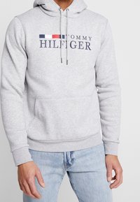 Sudadera gris de mezcla de algodón, con un bolsillo frontal, capucha con cordón y logo de "TOMMY HILFIGER" en color marino con rayas rojas y azules.