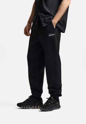 Mężczyzna ubrany w czarne spodnie dresowe z białym logo na udzie i czarne buty sportowe, stoi z jedną ręką w kieszeni.