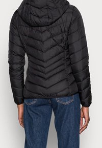 Černá péřová bunda s kapucí, s prošitým vzorem ve tvaru šipky, měkkou texturou a přiléhavým střihem, nošená přes modré džínové kalhoty.