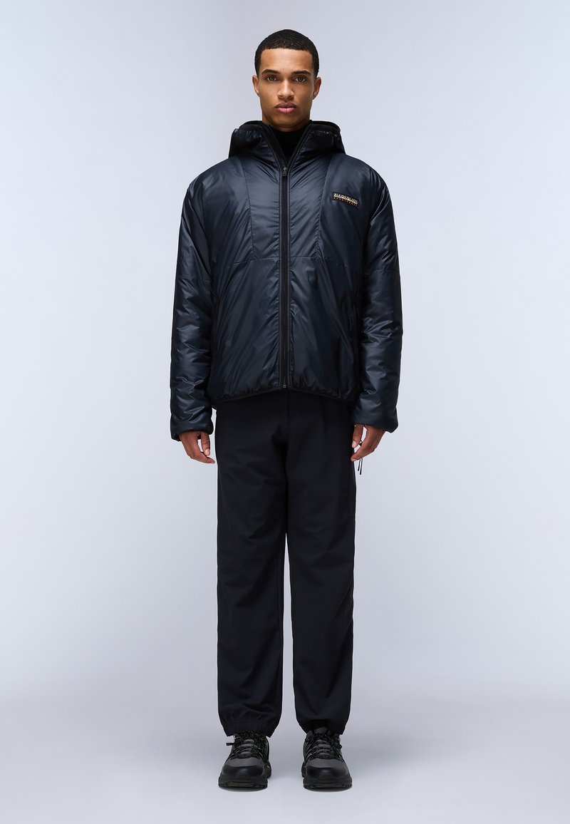 Czarna ocieplana kurtka z kapturem, połyskująca, z przednim zamkiem błyskawicznym i detalem logo na piersi. Noszona z ciemnymi spodniami i solidnymi butami.