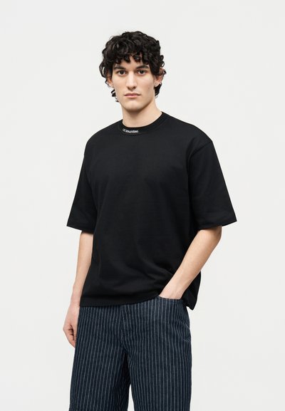 Νέος άνδρας με σγουρά μαλλιά, φοράει μαύρο φαρδύ t-shirt και σκούρο μπλε παντελόνι με κάθετες ρίγες, στέκεται με το ένα χέρι στη τσέπη.