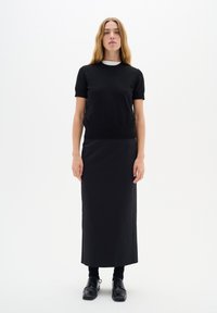 Musta slim fit lyhythihainen paita pyöreällä kaula-aukolla yhdistettynä pitkään mustaan hameeseen, molemmat valmistettu sileästä kankaasta, ja asusteena mustat kengät.