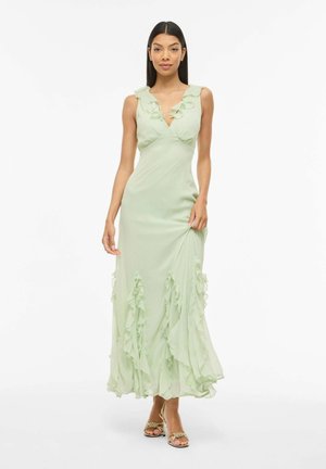 Kvinde iført en ærmeløs lys grøn maxikjole med flæser ved V-halsudskæringen og kanten, kombineret med guld åbne tå-hæle, stående mod hvid baggrund.