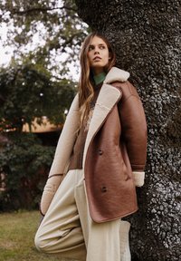 Brah bež usnjen plašč s kremasto ovčjo krzno ovratnikom, v kombinaciji z rjavim puloverjem in teksturiranimi bež hlačami, postavljenimi pred drevesnim ozadjem.