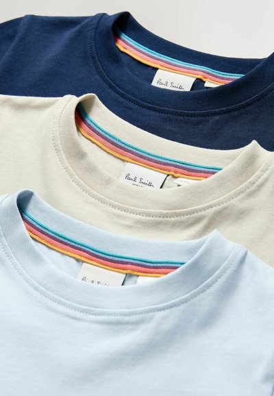 Drei gestapelte Baumwoll-T-Shirts: marineblau, beige und hellblau. Jedes hat einen farbenfrohen gestreiften Kragenbund und zeigt ein Markenetikett auf der Rückseite.