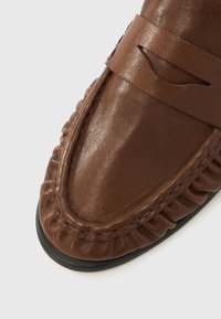 Καφέ δερμάτινα μοκασίνια με λεία υφή, στρογγυλερή μύτη, διακοσμητική ραφή και μάζεμα λεπτομέ ρειας γύρω από την άκρη. Σόλα με χαμηλό προφίλ.