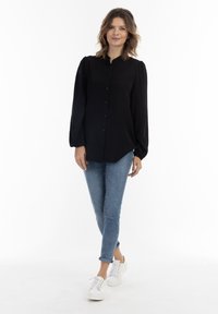 Zwarte button-up blouse met hoge kraag en puffmouwen, gecombineerd met lichtblauwe jeans en witte sneakers. Eenvoudig, schoon ontwerp.