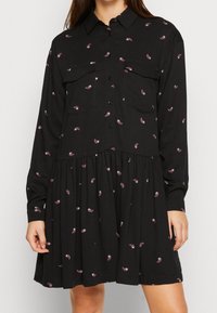 Svart klänning med lång ärm och krage, med två framfickor och ett flerlagerskjol, utsmyckad med små rosa paisleymönster.