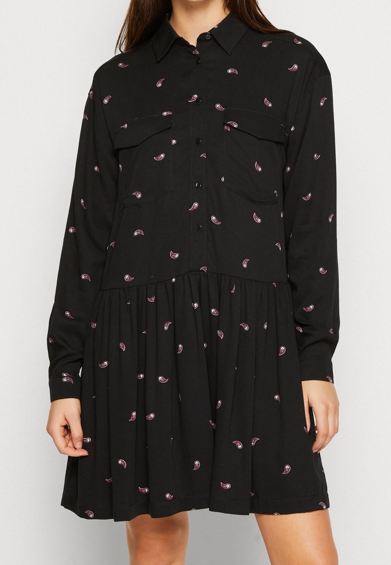 Svart klänning med lång ärm och krage, med två framfickor och ett flerlagerskjol, utsmyckad med små rosa paisleymönster.