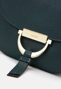 Sac à main en cuir teal avec un accent en métal doré comprenant un anneau circulaire et un fermoir rectangulaire, avec des coutures détaillées sur les bords.