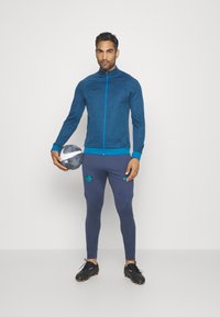 Herrenblauer Nike Reißverschlussjacke aus strukturiertem Stoff, kombiniert mit taillierten navy Hosen, die ein grafisches Logo und dekorative Nähte aufweisen. Schwarze Stiefel.