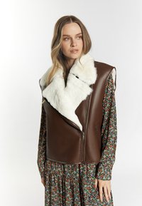 Bruine leren vest met een witte faux fur kraag en rits, gelaagd over een bloemenpatroon jurk met lange mouwen en een geplooide rok.