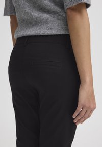 Čierne nohavice s elegantným dizajnom, ktoré majú hladkú textúru a dve zadné vrecká. Sivý pletený sveter čiastočne zakrýva pás.