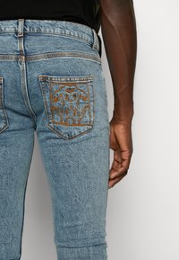 Jeans de denim azul claro con corte ajustado. Cuenta con un diseño bordado en naranja en el bolsillo trasero. Costuras visibles a lo largo de las costuras.