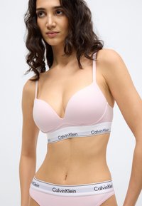 Ροζ ανοιχτό ροζ μπουστάκι με βαθύ ντεκολτέ και λεπτές τιράντες, με λευκή ταινία με το λογότυπο "Calvin Klein". Λείο ύφασμα και καμπυλωτό σχήμα.