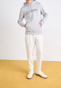 Grå huva sweatshirt med svart AMG-logotyp, ihopparad med vita byxor med sidofickor med dragkedja. Sportiga skor med bruna accenter fulländar outfiten.