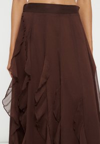 Jupe marron foncé avec des panneaux de tissu superposés et froncés, visible de la taille jusqu’au milieu de la cuisse, portée par une personne avec le ventre découvert et les bras visibles.