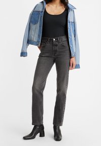 Jeans negros de tiro alto descoloridos, top negro sin mangas con relieve y chaqueta de mezclilla azul clara con bolsillos. Textura suave, corte recto y botas de tacón.