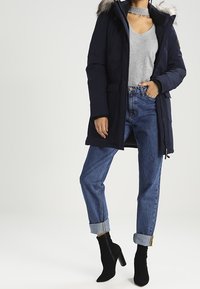 Marinblå pufferjacka med fuskpäls, grå topp, hög midja blå jeans som är uppvikt i ärmarna, och svarta ankelskor. Prickfritt sytt.