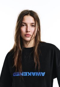 Hanorac negru supradimensionat din bumbac, cu un text grafic albastru inversat "AWAKENED" pe piept. Păr natural care acoperă parțial fața.