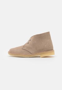 Stivale alla caviglia in suede beige con punta arrotondata, due occhielli per i lacci e una leggera suola in gomma. Texture liscia con sottili dettagli di cucitura.