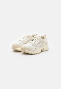 Lys beige treningssko med netting og syntetiske materialer, rund tå, polstret såle og subtile mønstrede detaljer.