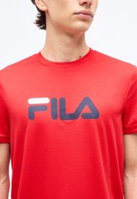 Νέος άνθρωπος που φοράει φωτεινό κόκκινο μπλουζάκι με το λογότυπο FILA, κομμένο λίγο κάτω από το πηγούνι, σε απλό λευκό φόντο.