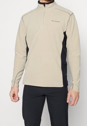 Mand iført en beige og sort Columbia fleece pullover med kvart lynlås og sorte bukser mod en ensfarvet hvid baggrund.