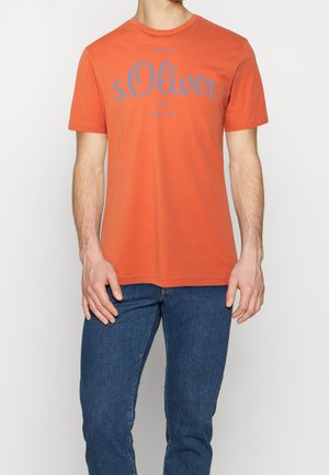 Mand iført orange kortærmet T-shirt med blå teksten "s.Oliver" og mørkeblå jeans, stående foran en ensfarvet baggrund.
