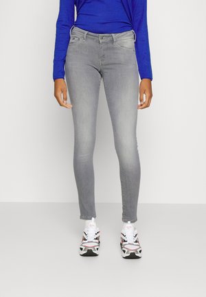 Calças jeans skinny cinzentas com cinco bolsos, fecho de braguilha e passantes para cinto, combinadas com sapatilhas brancas e uma blusa azul de mangas compridas.