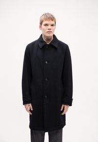 Cappotto di lana nera con colletto, chiusura frontale a sei bottoni e tasche a patta. Presenta un fit su misura e una silhouette strutturata.
