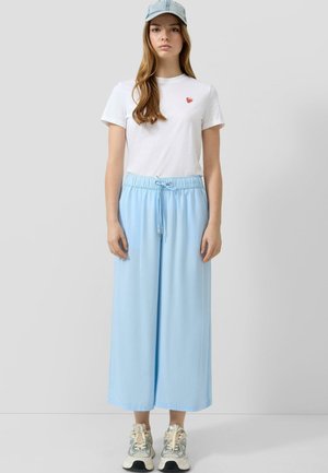 Młoda kobieta ubrana w białą koszulkę z małym czerwonym sercem, jasnoniebieskie szerokie spodnie ze sznurkiem, trampki i jasnoniebieską czapkę z daszkiem.