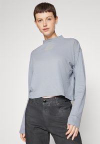 Bluză scurtă cu mânecă lungă din material albastru deschis, având un guler înalt și un logo brodat subtil. Asortată cu pantaloni gri închis.