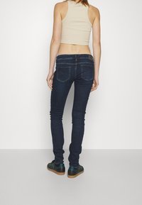 Jeans ajustados azul oscuro con una cintura de media altura, que cuentan con un yugo trasero y dos bolsillos traseros, combinados con un top corto beige y zapatillas verdes.