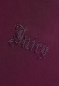 Σκούρο μωβ ύφασμα με τη λέξη "Juicy" γραμμένη με διακοσμητική γραφή χρησιμοποιώντας μικρά λαμπερά στρας.