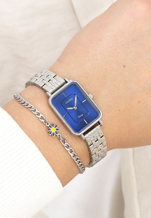 Montre en acier argenté avec un cadran bleu rectangulaire, des index argentés et un bracelet en acier inoxydable. Accompagnée d'un bracelet en chaîne argenté avec un charme en forme de marguerite.