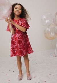 Abito rosso con paillettes e maniche arricciate, con motivo a spirale rosa. La bambina indossa scarpe rosa scintillanti e tiene in mano un palloncino trasparente.