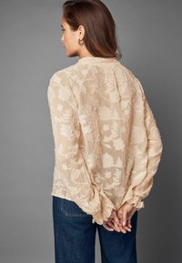 Blouse beige à motif floral en tissu transparent, avec des manches bouffantes et des poignets froncés, associée à un jean bleu foncé. Vue de dos.