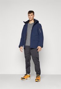 Marine isolert jakke med hette, flere lommer og grå fôr, kombinert med grå cargo bukser og oransje joggesko.
