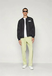 Black bomber jacket with logo on chest, white t-shirt, light green trousers, and white sneakers. Model wearing sunglasses, posing against a neutral background.