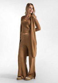 Bruin gebreid loungewear-set met een aansluitend topje, wijde broek en een lange cardigan met zijslits; zachte textuur en warme tinten.