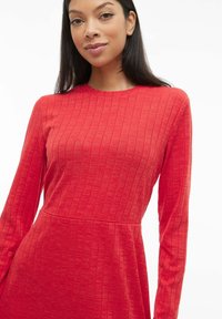 Robe rouge côtelée à manches longues, col rond et jupe évasée. Confectionnée dans un tissu doux et texturé avec de fines lignes verticales.