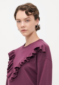 Giovane donna con capelli corti e ondulati indossa orecchini a cerchio e una maglietta viola con dettagli a volant diagonali sul petto e sulla spalla.